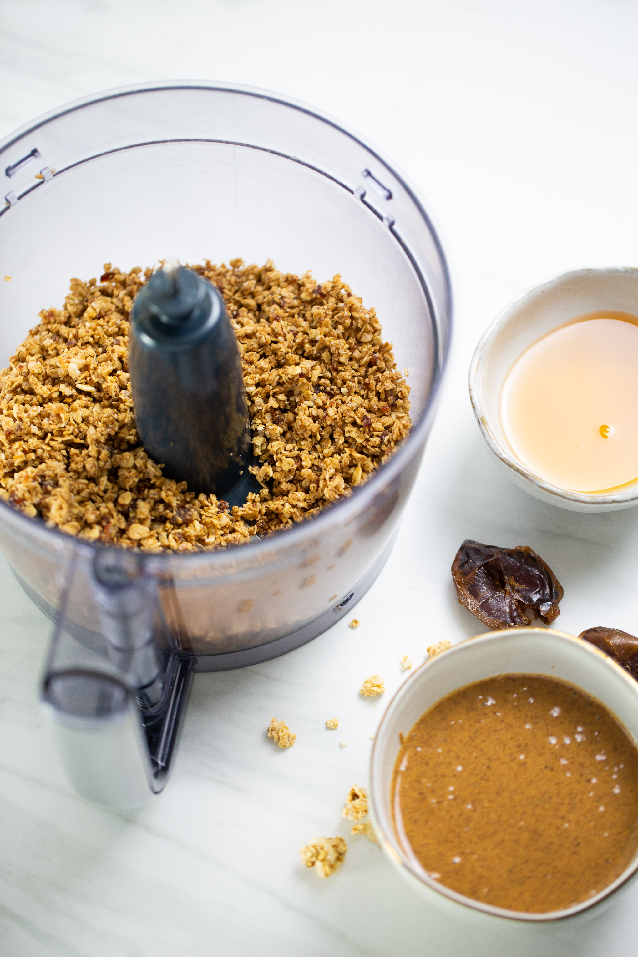 Food processor bowl with granola, a bowl of almond butter and another ewith maple syrup on the side.