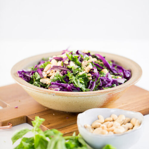 A bowl filled with vibrant Asian red cabbage salad—featuring shredded purple cabbage, chopped herbs, and cashews—sits on a wooden board. In the foreground, a small bowl holds more cashews next to fresh basil leaves, all set against a white background.