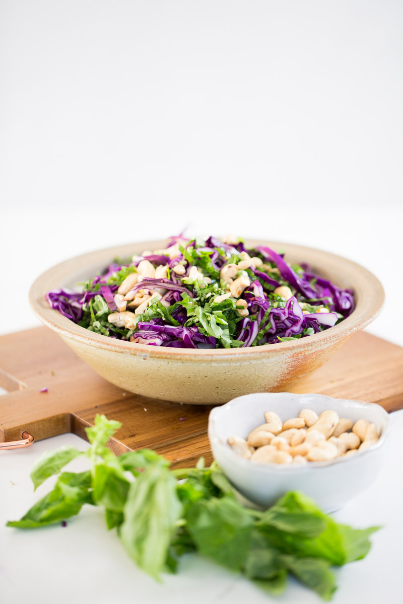 Asian red cabbage salad in a bowl, nex to a bowl of cashews and basil.