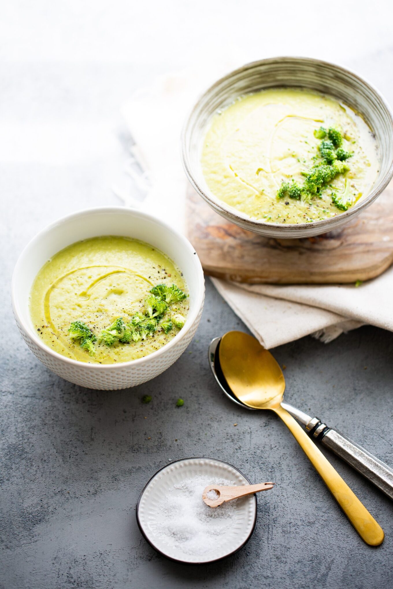Two bowls of creamy vegan broccoli soup are shown; one is on a wooden board, the other on a gray surface. Both are garnished with herbs and olive oil, with two spoons, a folded napkin, and coarse salt in a small dish nearby.