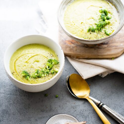 Two bowls of creamy vegan broccoli soup are shown; one is on a wooden board, the other on a gray surface. Both are garnished with herbs and olive oil, with two spoons, a folded napkin, and coarse salt in a small dish nearby.