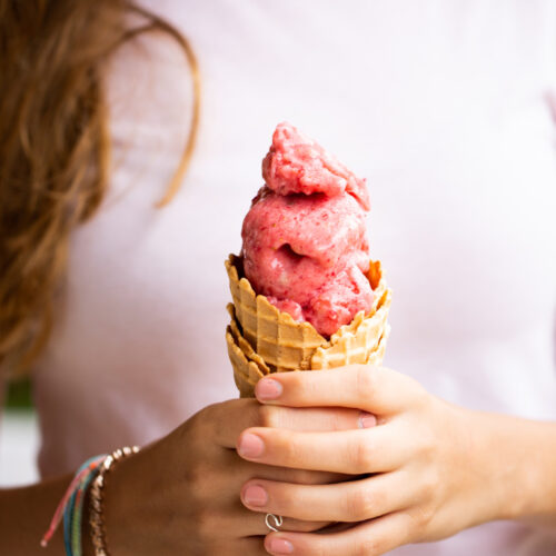 A person in a white shirt holds a waffle cone brimming with three scoops of creamy, pink banana strawberry blender ice cream. Their long brown hair cascades down as colorful bracelets and a ring adorn their hands. The focus remains on the delightful treat, set against a softly blurred background.
