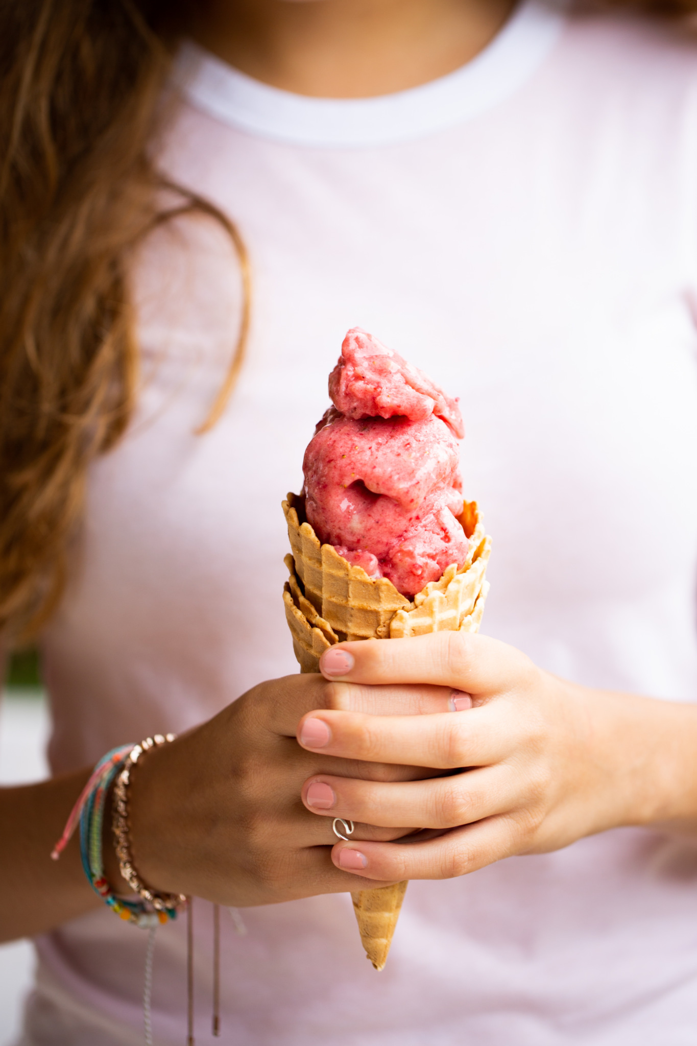 A person in a white shirt holds a waffle cone brimming with three scoops of creamy, pink banana strawberry blender ice cream. Their long brown hair cascades down as colorful bracelets and a ring adorn their hands. The focus remains on the delightful treat, set against a softly blurred background.