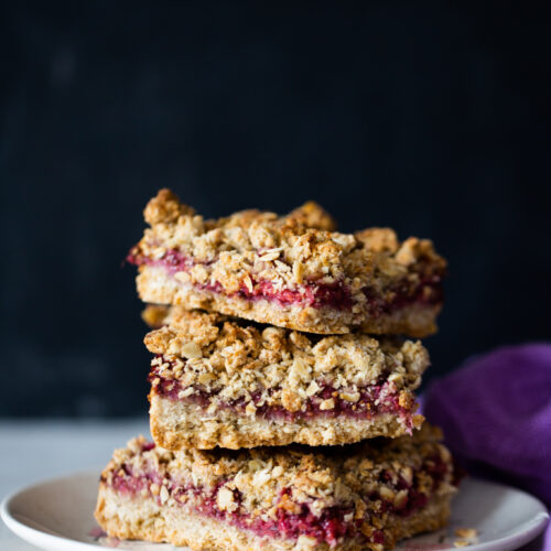 Three stacked vegan oat bars with berry crumble sit on a white plate against a dark background. The bars feature visible layers of oats, crumble topping, and thick, red berry filling. A purple cloth is partially visible to the right of the plate.
