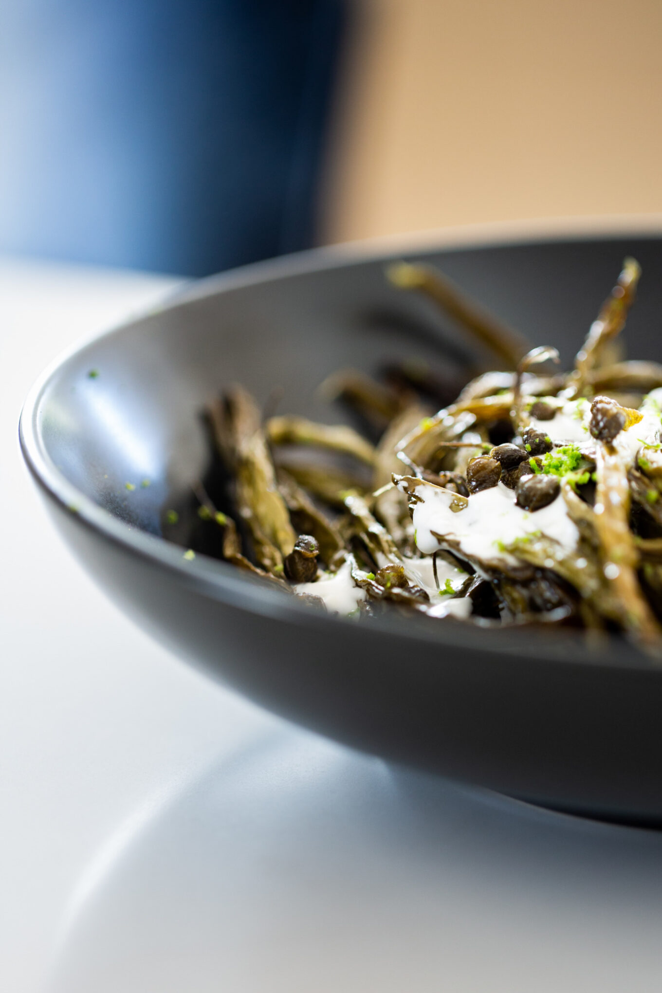 A black bowl filled with baked green beans topped with a creamy white sauce and sprinkled with capers. The dish is garnished with lime zest and fresh herbs. The shallow depth of field creates a blurred background, highlighting the texture and details of the ingredients.
