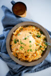 A brown bowl of creamy mashed potatoes topped with a generous dollop of cheese sauce and garnished with chopped parsley. The mashed potatoes are made from sweet potato for added flavor. The bowl sits on a crumpled blue cloth near a small black cup filled with vegan chipotle queso, all on a white surface.