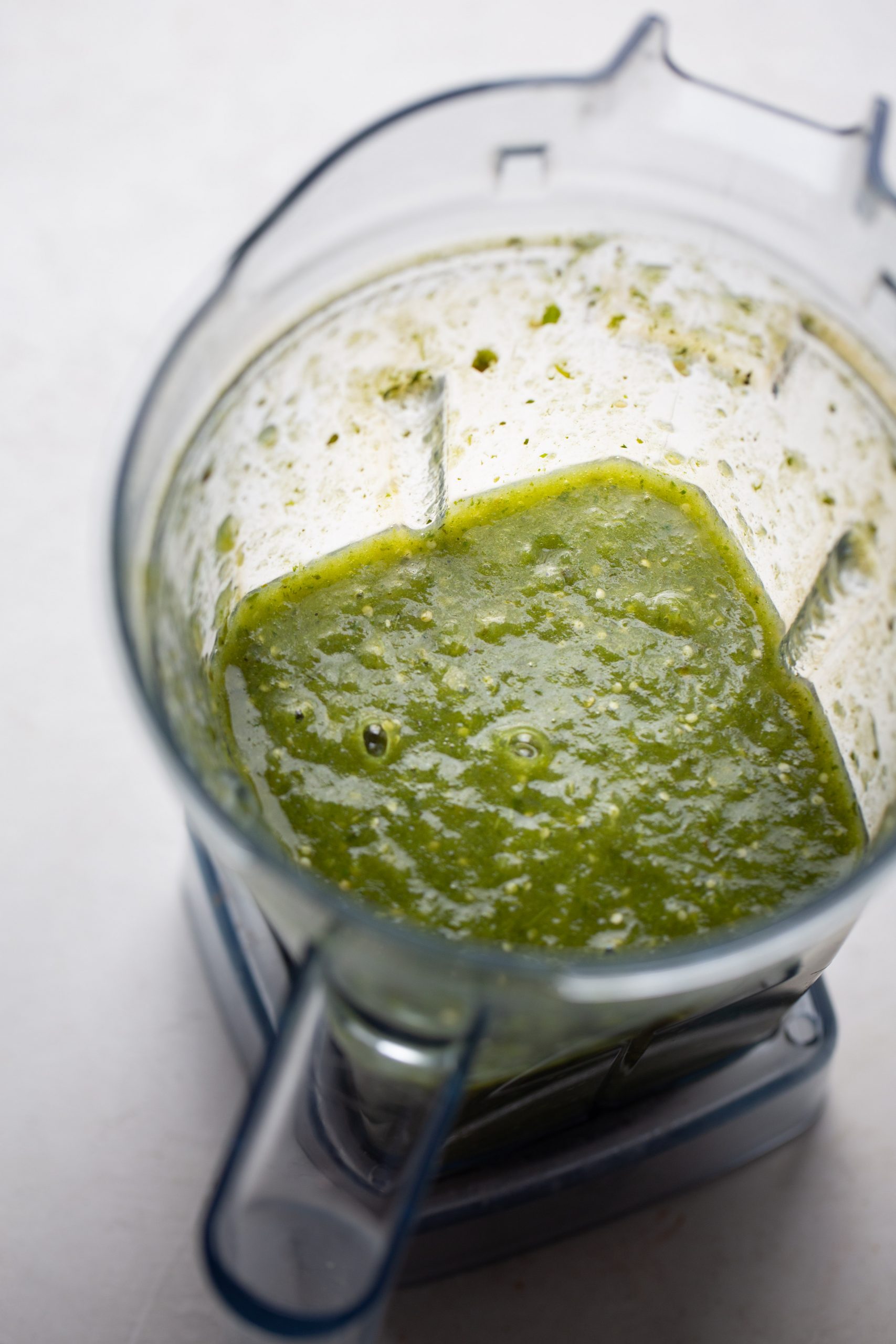 A clear blender pitcher containing a thick, vibrant green sauce for no-bake enchiladas sits on a light surface. The slightly chunky mixture clings to the sides, showing it has recently been blended.