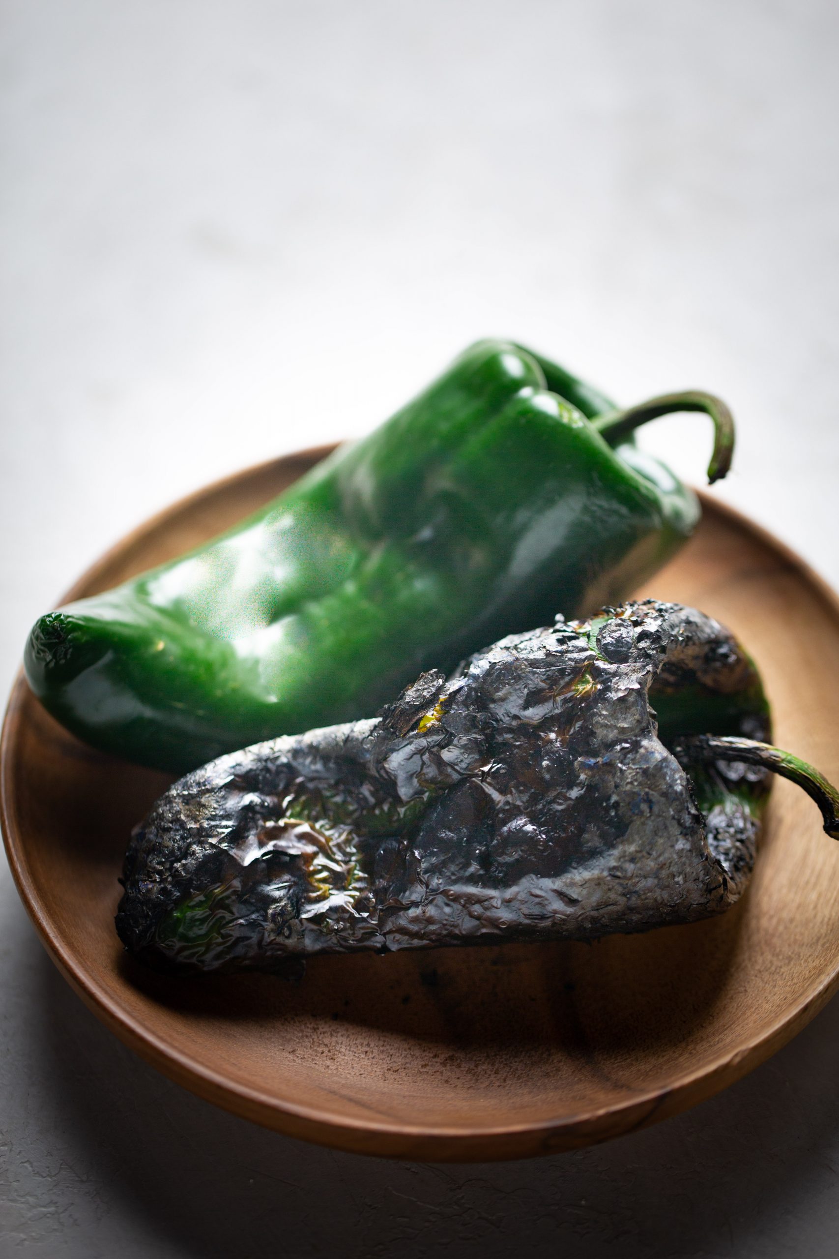 A round wooden plate holds two poblanos—one fresh and green, the other charred with blackened, blistered skin—perfect for adding depth to no-bake enchiladas. The background is a light, neutral surface.