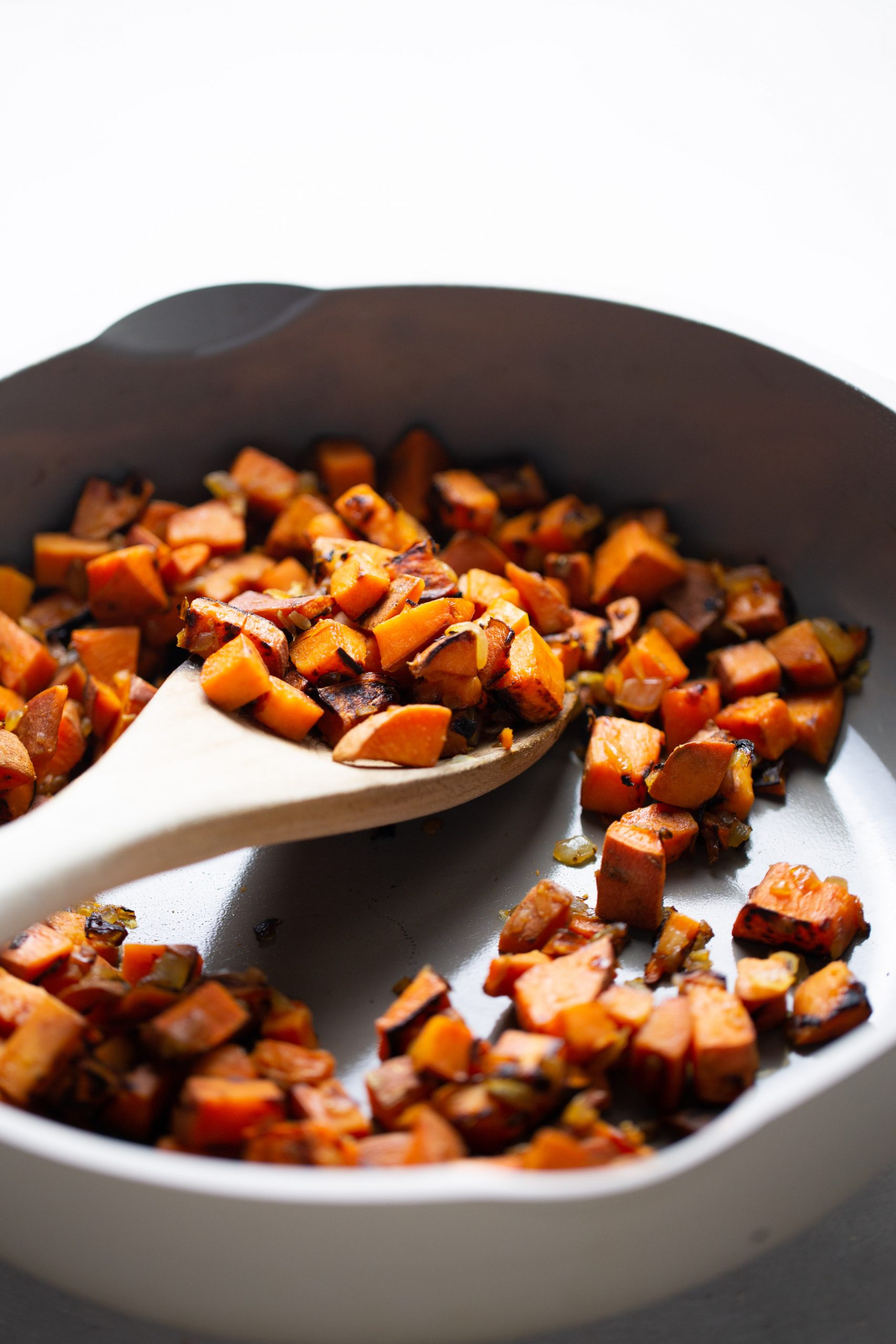 A white skillet contains sautéed, cubed sweet potatoes with caramelized edges—perfect for adding to no-bake enchiladas. A wooden spoon lifts the crisp, orange cubes. The bright background is softly out of focus.