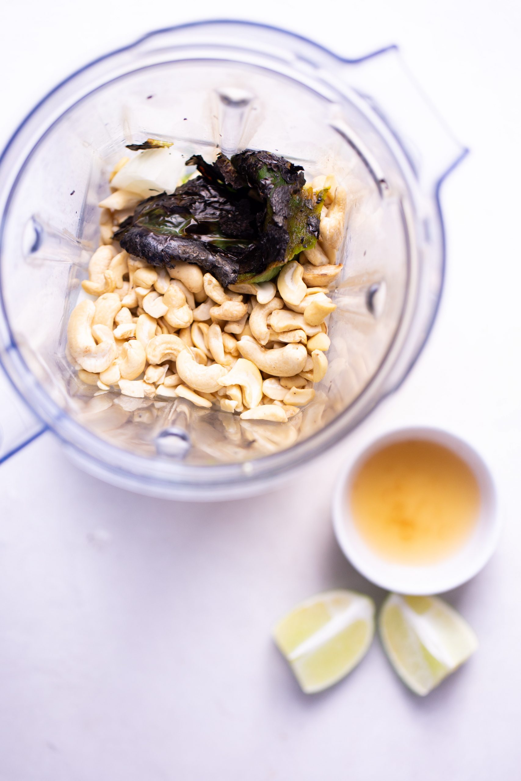 A blender holds raw cashews, charred poblano pepper, and a small piece of onion—the perfect base for a creamy no-bake enchiladas sauce. Nearby sit a white bowl with light brown liquid and two lime wedges on a white surface.