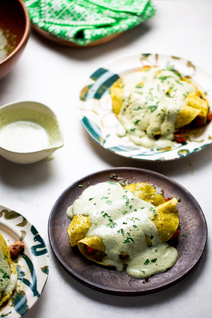 Dos platos con enchiladas de camote doradas y dobladas, generosamente cubiertas de cremosa salsa verde y espolvoreadas con hierbas frescas. Un pequeño cuenco con salsa y una servilleta con motivos verdes están cerca, sobre una superficie clara. La comida parece fresca y vibrante.