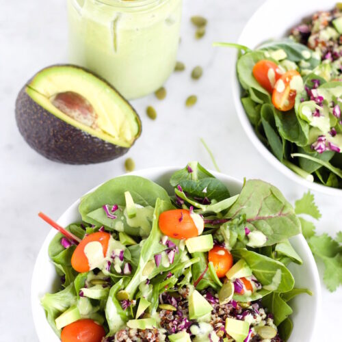 Two bowls filled with a colorful salad featuring spinach, cherry tomatoes, diced avocado, quinoa, and red cabbage are drizzled with creamy cilantro mint dressing. A halved avocado and a jar of the vibrant green dressing with a wooden spoon lie in the background on a white surface.