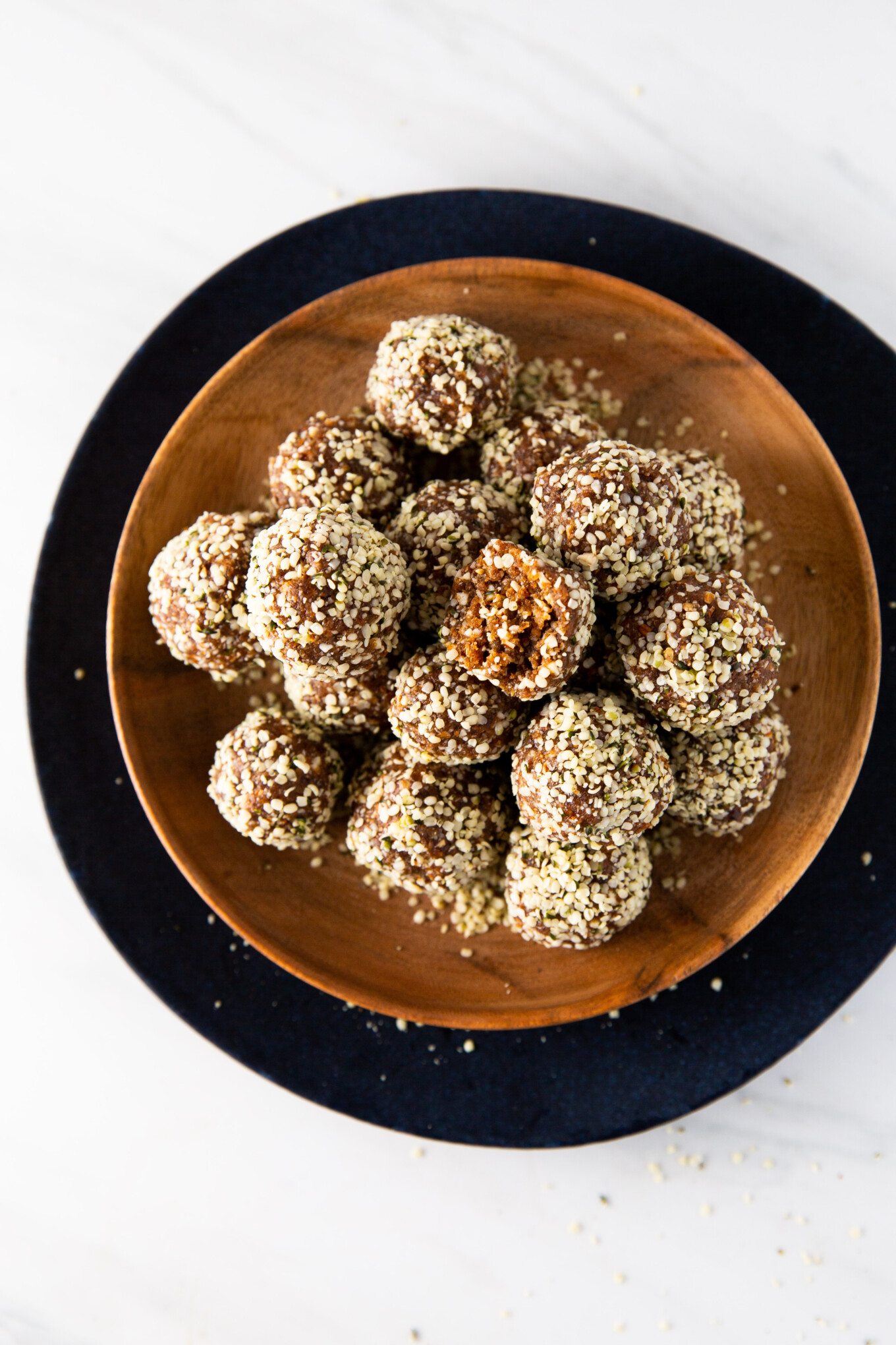 A wooden bowl on a dark round plate holds several vegan protein balls covered in hemp seeds. One bite has a small piece missing, revealing a chewy texture. The background is a light marble surface.