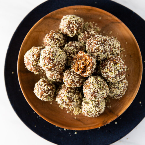 A wooden bowl on a dark round plate holds several vegan protein balls covered in hemp seeds. One bite has a small piece missing, revealing a chewy texture. The background is a light marble surface.