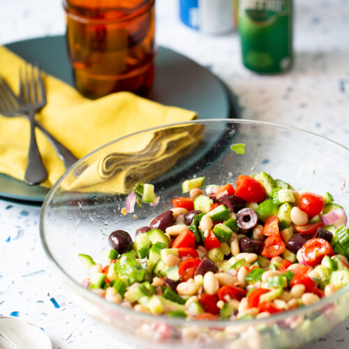 A glass bowl filled with a colorful ensalada mediterranea—bursting with tomatoes, cucumbers, green peppers, olives, and beans—rests on a terrazzo surface. Beside it lie a metal spoon, a plate of folded yellow napkins, utensils, amber glasses, and beverage cans.