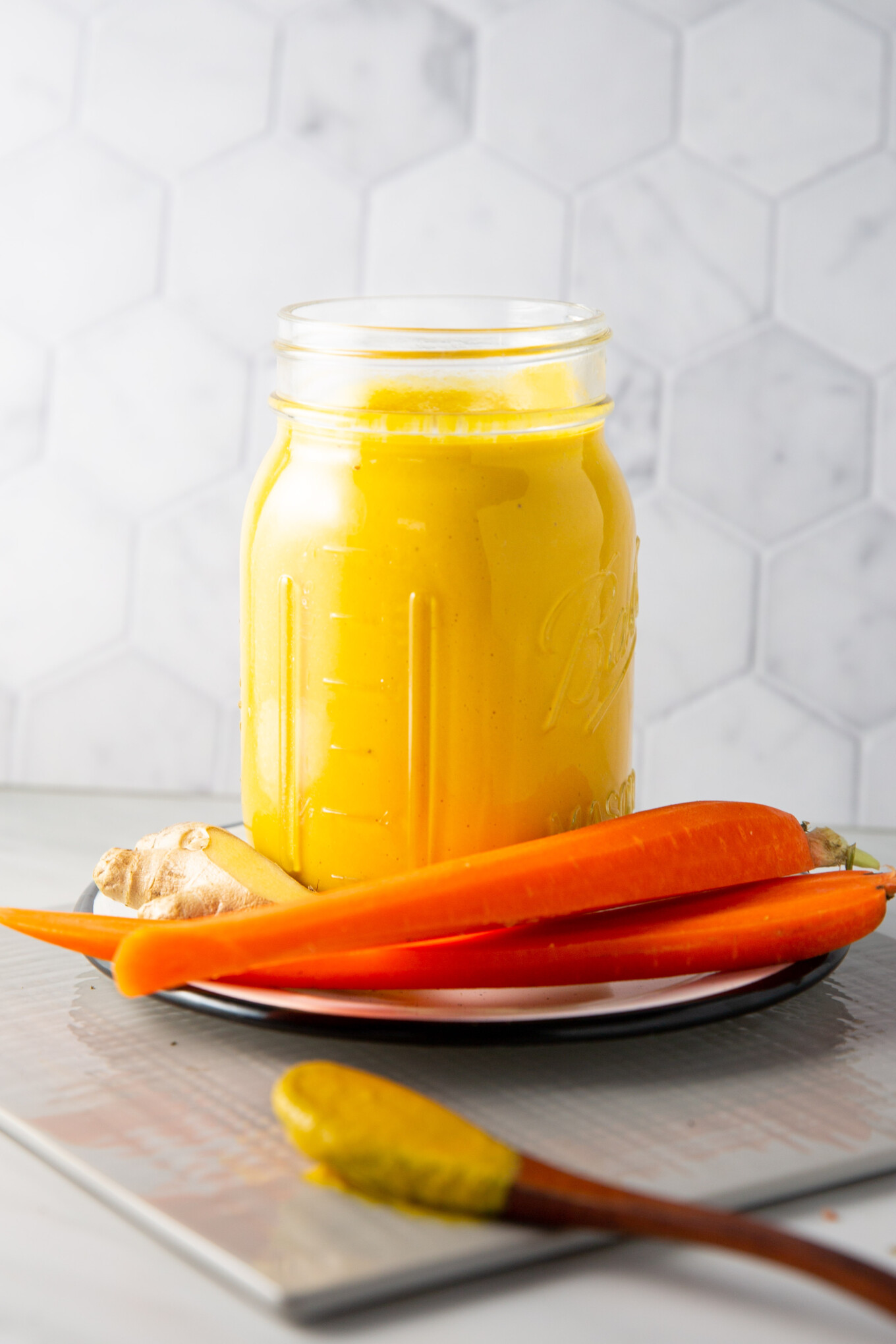A mason jar filled with bright yellow carrot ginger dressing sits on a plate. The plate also holds three fresh carrots and a piece of ginger. A wooden spoon with some of the perfect yellow dressing on its tip rests in the foreground. The background features a hexagonal tile pattern.