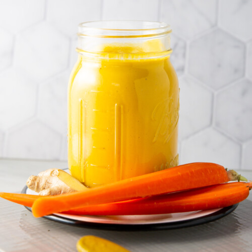 A mason jar filled with bright yellow carrot ginger dressing sits on a plate. The plate also holds three fresh carrots and a piece of ginger. A wooden spoon with some of the perfect yellow dressing on its tip rests in the foreground. The background features a hexagonal tile pattern.