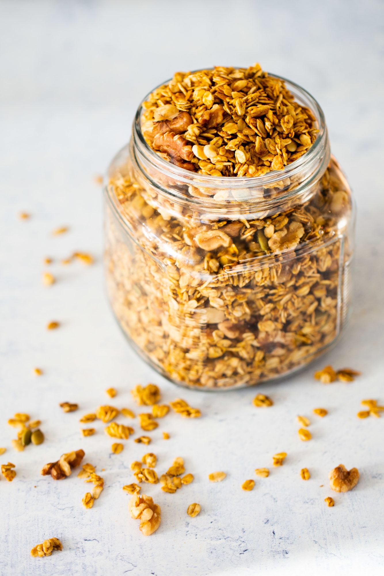 A glass jar filled with pumpkin pie granola, featuring rolled oats, nuts, and seeds. Scattered granola pieces add a casual, rustic touch on the light surface. The background is softly blurred in white and light blue tones, enhancing the cozy autumn vibe.