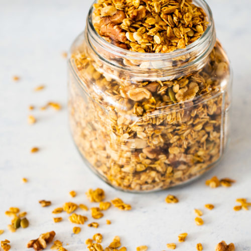 A glass jar filled with pumpkin pie granola, featuring rolled oats, nuts, and seeds. Scattered granola pieces add a casual, rustic touch on the light surface. The background is softly blurred in white and light blue tones, enhancing the cozy autumn vibe.
