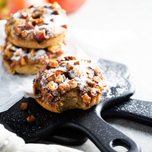 Close-up of apple cinnamon muffins with caramelized apple chunks, reminiscent of baked apple donuts, dusted with powdered sugar on black slate paddles. A bite is taken from the front muffin. Two red apples are blurred in the background, and a white cloth is partially visible in the foreground.