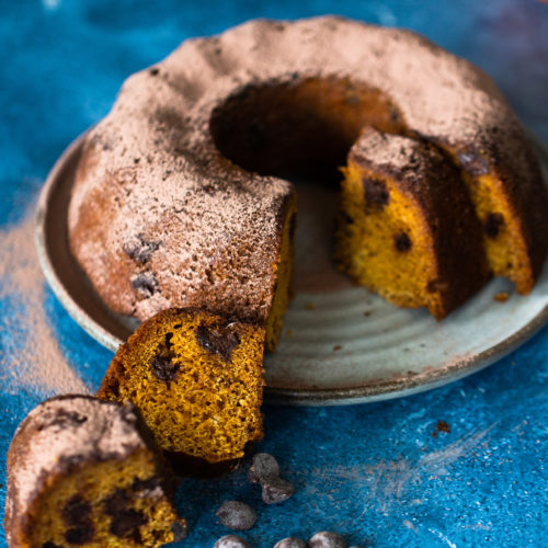A round vegan chocolate chip bundt cake is on a plate, topped with a dusting of cocoa powder. Slices are cut, revealing a golden interior with visible chocolate chips. A few chocolate chips are scattered on the blue surface beside the plate. An orange fruit is blurred in the background.