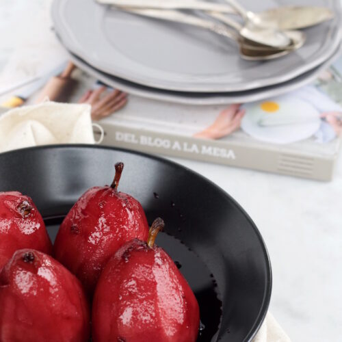 A black bowl contains four glossy red wine poached pears. They are elegantly arranged on a beige cloth with a cookbook peeking beneath. In the background, stacked gray plates with silver cutlery rest atop a light-colored surface, completing the refined setting.