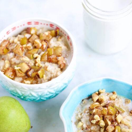 Two bowls of oats and amaranth porridge topped with diced pears, walnuts, and syrup sit on a light surface. Nearby are a whole pear, measuring spoons with spices, and a jar of milk. The patterned bowls add a charming touch.
