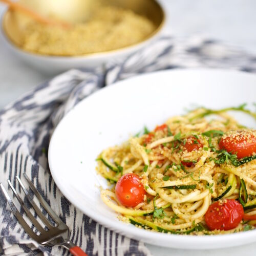 A white bowl of spaghetti and zucchini noodles with cherry tomatoes, herbs, and breadcrumbs sits on a patterned cloth next to a fork. In the background, there’s a golden bowl of extra breadcrumbs and a small wooden spoon on a marble surface.
