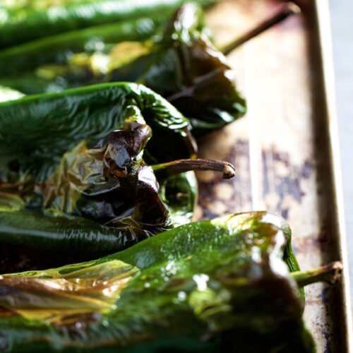 A close-up of roasted poblano peppers on a metal baking tray reveals their blistered skins with charred spots, showcasing the roasting process. The vibrant green color contrasts with darker, browned portions. The background features a dark, out-of-focus surface, highlighting the peppers' texture.