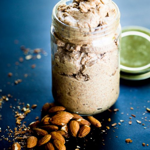 A jar of homemade almond butter sits open on a dark surface. Whole almonds are scattered nearby, with the lid partially visible in the background. Crumbs and almond pieces are spread around, enhancing the rustic and authentic feel.