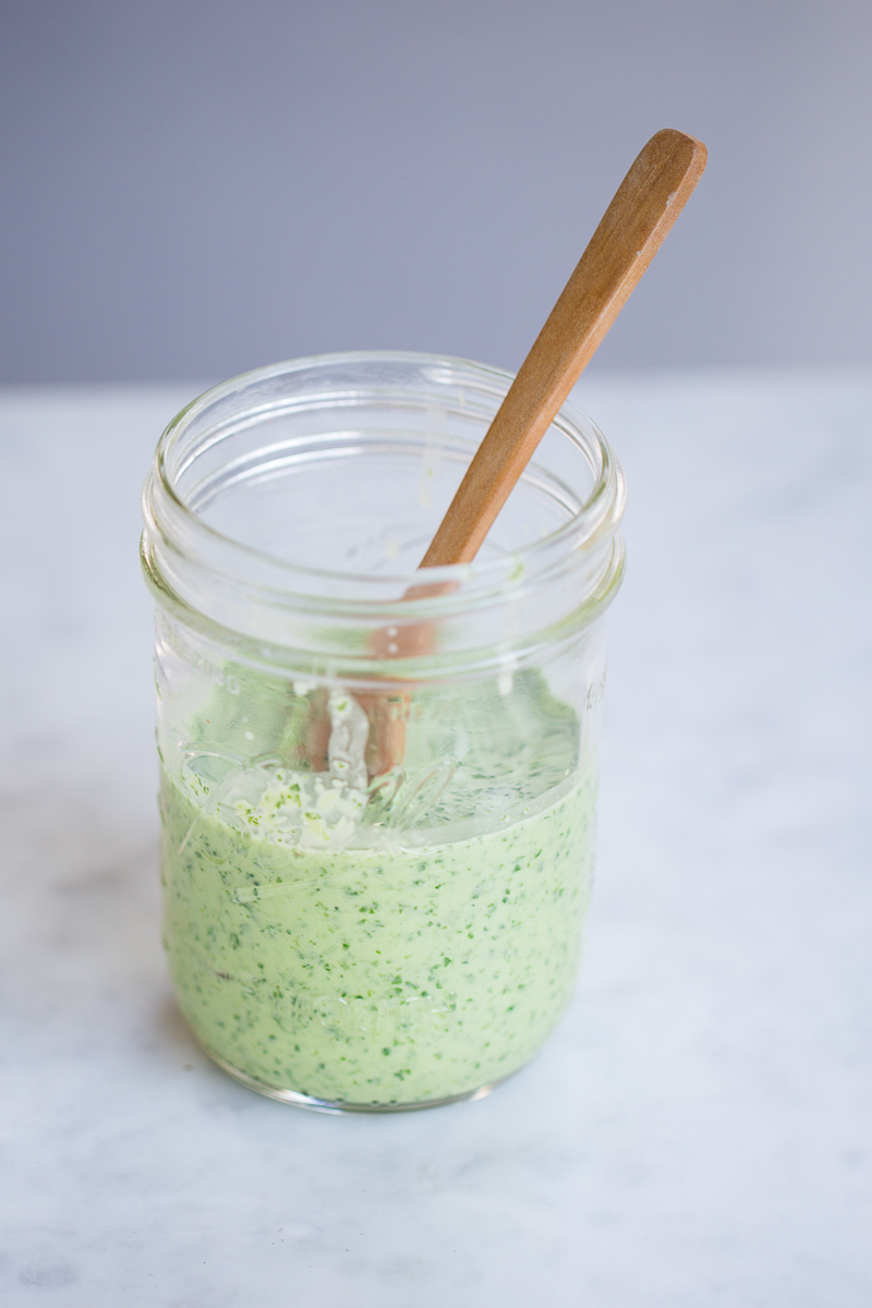 A glass jar filled with creamy, pale green cilantro dressing flecked with herbs sits on a white marble surface. A wooden spoon rests inside the jar, its handle leaning out. The background is softly blurred in shades of gray and blue.