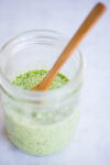 A clear glass jar filled with creamy, green cilantro dressing speckled with herbs sits on a pale marble surface. A wooden spoon rests inside the jar, partially submerged in the mixture. The background is softly blurred.