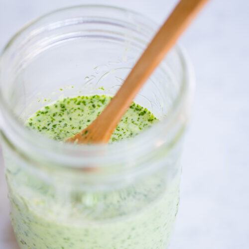 A clear glass jar filled with creamy, green cilantro dressing speckled with herbs sits on a pale marble surface. A wooden spoon rests inside the jar, partially submerged in the mixture. The background is softly blurred.