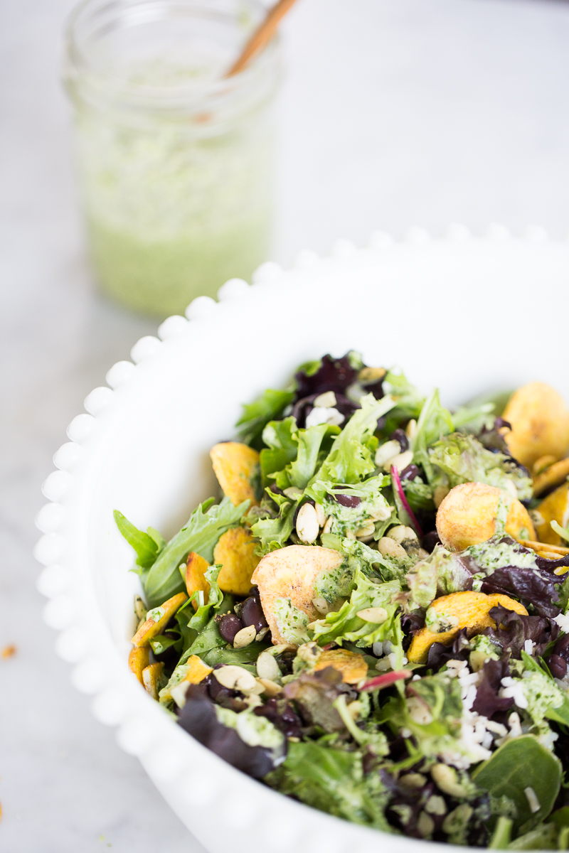 A close-up of a fresh green salad in a white bowl, topped with sliced almonds, seeds, plantain chips, and a creamy cilantro dressing. In the background, a glass jar filled with more green dressing and a wooden spoon is slightly out of focus.