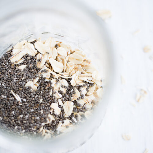A close-up, top-down view of a glass jar filled with oats and chia, perfect for an overnight oats and chia breakfast, sits on a white surface with scattered loose oats. Soft natural lighting highlights the bright and wholesome scene.