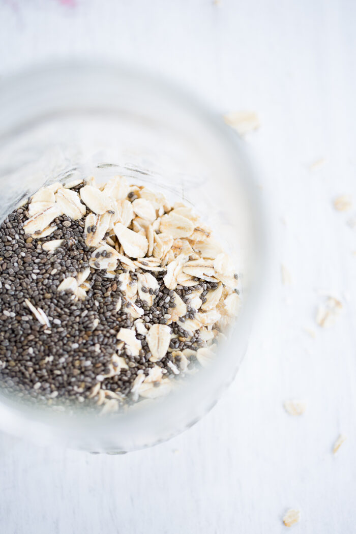 A close-up, top-down view of a glass jar filled with oats and chia, perfect for an overnight oats and chia breakfast, sits on a white surface with scattered loose oats. Soft natural lighting highlights the bright and wholesome scene.