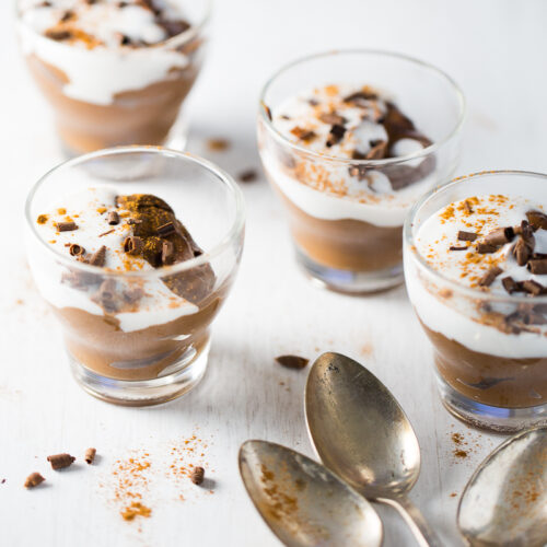 Four glass cups filled with avocado chocolate pudding, topped with whipped cream, chocolate shavings, and a sprinkle of spices, are placed on a white surface. Three tarnished silver spoons rest in the foreground beside the desserts.
