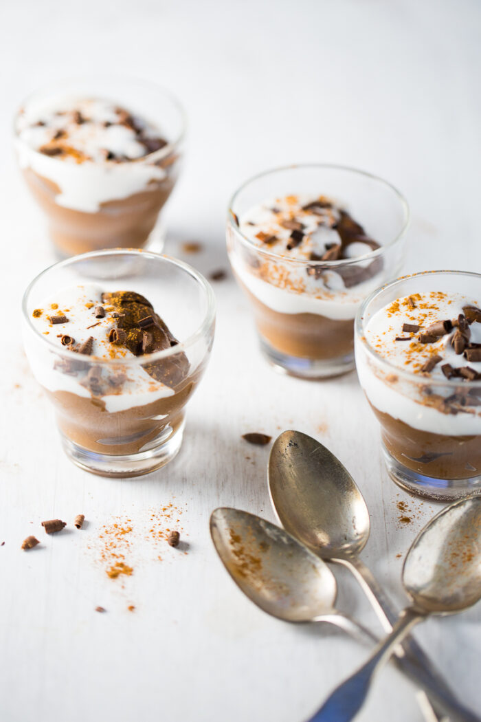 Four glass cups filled with avocado chocolate pudding, topped with whipped cream, chocolate shavings, and a sprinkle of spices, are placed on a white surface. Three tarnished silver spoons rest in the foreground beside the desserts.