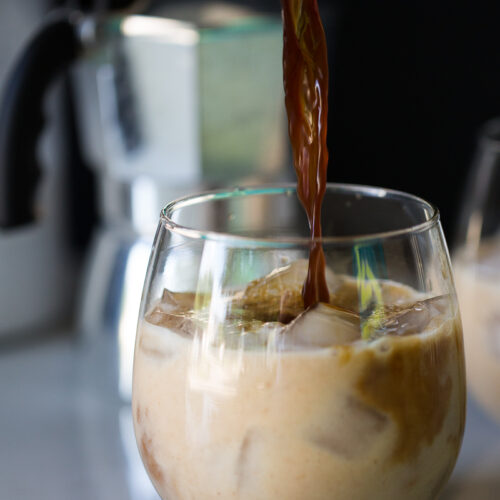 A glass filled with iced milk is being poured over with coffee, creating a swirl of mixed colors. The background features a blurred silver espresso maker with a black handle. The surface is a light-colored countertop.