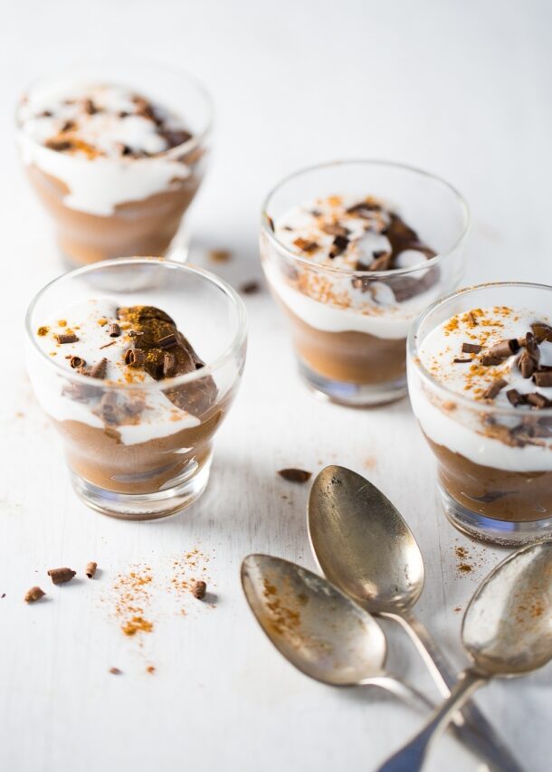 Cuatro vasos de cristal llenos de cremoso pudín de chocolate con aguacate, cubierto con crema batida, virutas de chocolate y una pizca de polvo marrón, se disponen sobre una superficie blanca. Tres cucharas vintage se encuentran cerca, añadiendo un toque rústico al postre.