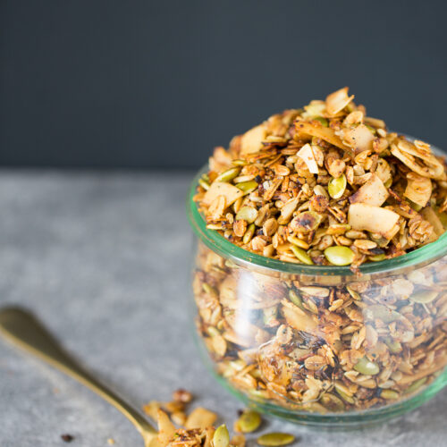 A glass jar filled with savory granola is placed on a gray surface. The granola contains oats, sliced almonds, pumpkin seeds, and various nuts. Beside the jar, a gold-colored spoon with some granola on it rests on the surface, with a few scattered pieces around it. The background is dark gray.