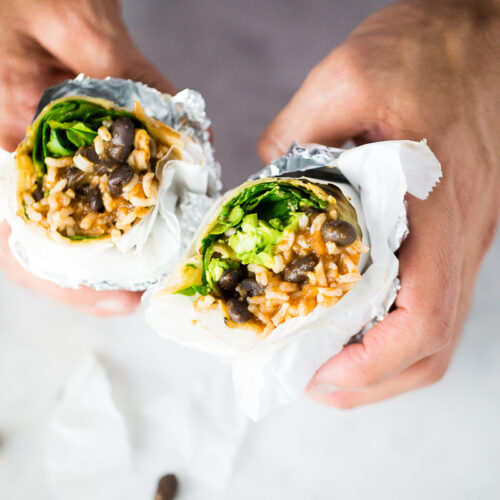 Two hands hold a divided veggie burrito wrapped in foil and paper. The burrito is filled with black beans, rice, greens, and other vegetables. A few black beans are scattered on a white surface below. The background is a slightly blurred light grey, highlighting the delicious allure of burritos.