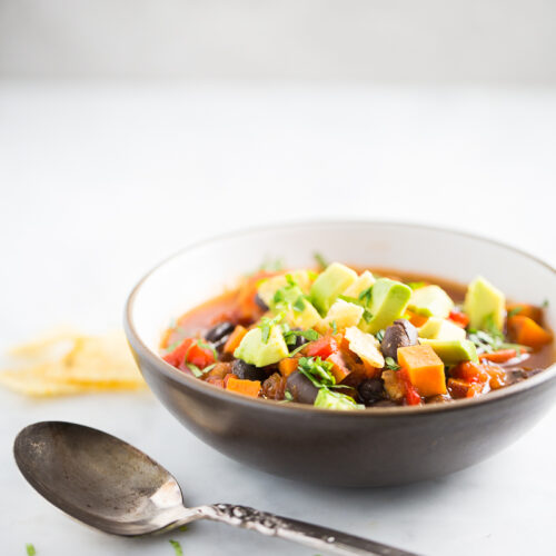A bowl of sweet potato chili topped with diced avocado and chopped herbs sits on a white surface. A vintage silver spoon lies in the foreground, and a tortilla chip is visible in the background. The scene is bright and minimalistic.