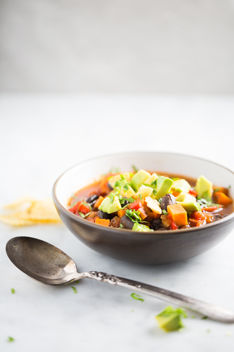 A bowl of sweet potato chili topped with diced avocado and chopped herbs sits on a white surface. A vintage silver spoon lies in the foreground, and a tortilla chip is visible in the background. The scene is bright and minimalistic.