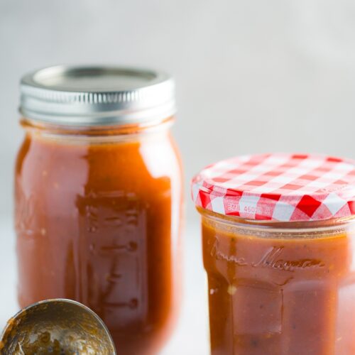 Two glass jars filled with Chipotle Enchilada Sauce are placed on a white countertop. The jar on the left has a metal lid, while the jar on the right has a checkered red and white lid labeled "Bonne Maman." A metal spoon with some sauce on it is in front of the jars. The background is light grey.