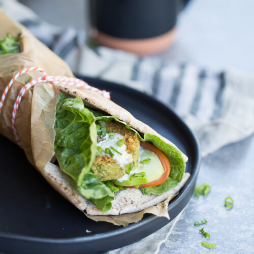 A wrapped falafel sandwich is placed on a black plate. It features lettuce, tomato slices, cucumber, and falafel inside a flatbread, partially bundled in brown paper and tied with red and white string. In the background, a black jug and striped cloth can be seen on a gray surface.