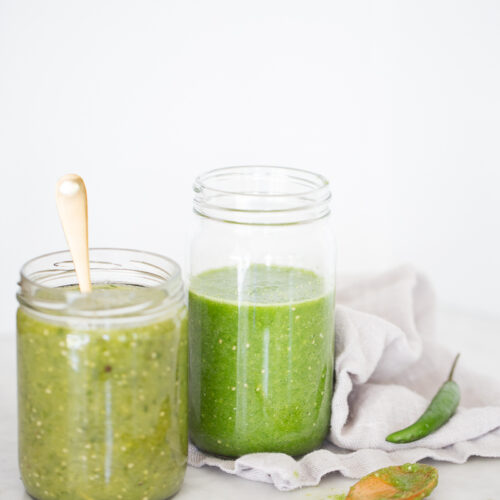 Two glass jars filled with green salsa verde sit on a light-colored marble surface. One jar, showcasing raw tomatillo salsa verde, has a wooden spoon inside, while the other jar is taller and lidless. Nearby, a wooden spoon with salsa and a green chili pepper rest on a light gray cloth against the plain white background.