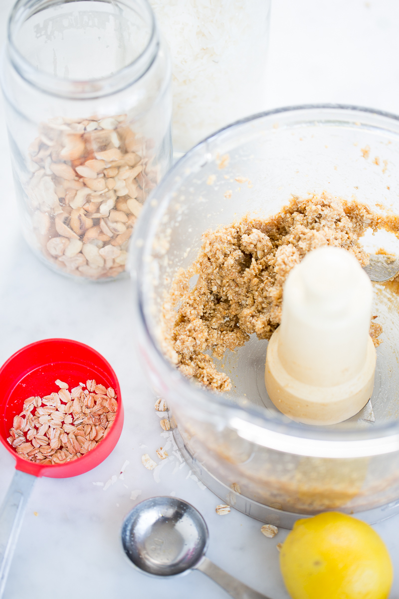 Sobre una encimera blanca hay un robot de cocina con una mezcla de avena y frutos secos. Cerca hay un tarro de frutos secos mezclados, coco rallado, avena en un vaso rojo, una cucharada de metal y un limón: ingredientes para hacer bolitas de limón caseras u otro sabroso tentempié.