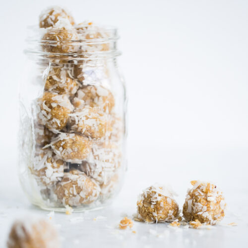 A glass jar filled with round, golden-brown coconut energy balls sits on a white surface. Three similar energy balls are outside the jar, some rolled in shredded coconut—perfect alongside overnight oats with chia for a wholesome snack.