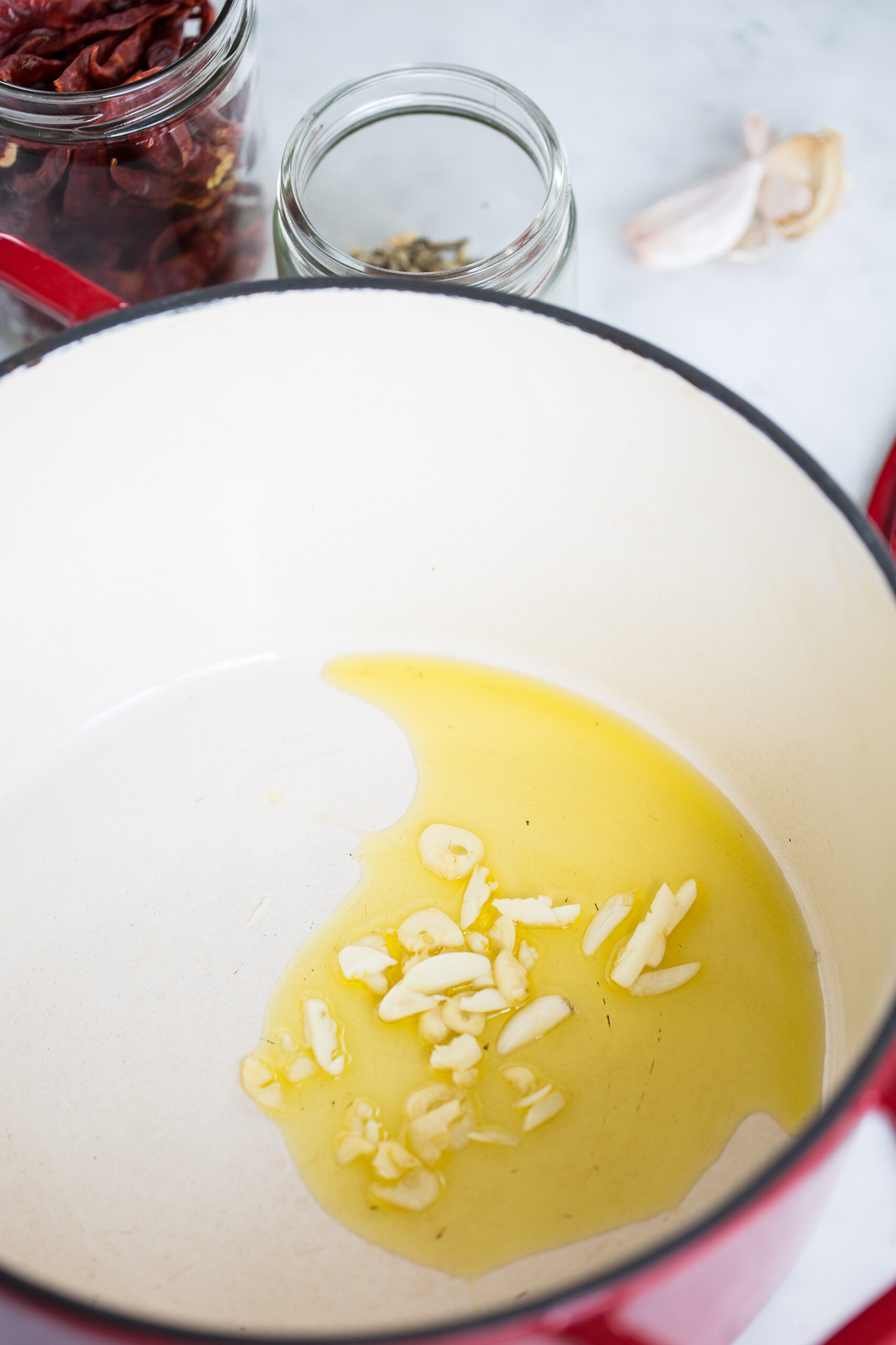 A white enameled pot with sliced garlic saut&eacute;ing in olive oil hints at the start of a one-pot pasta. Nearby, jars of dried red chilies and spices evoke the flavors of a classic puttanesca vegan dinner in the making.